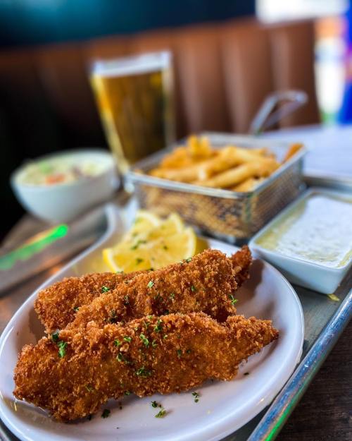 A plate of crispy fried fish fillets with lemon, coleslaw, fries, and dipping sauce, alongside a glass of beer.