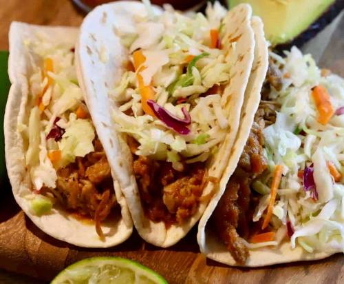 Three soft tacos filled with shredded meat and topped with colorful cabbage and carrot slaw, served on a wooden board.