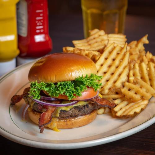 A juicy burger with bacon, lettuce, tomato, and cheese, served with crispy waffle fries and condiments in the background.