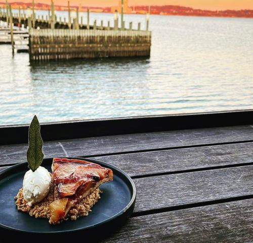 A slice of dessert with a scoop of ice cream on a black plate, overlooking a serene waterfront at sunset.