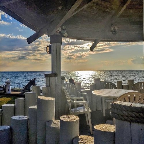 A seaside view from a gazebo, featuring a table and chairs, with the sun setting over the water.