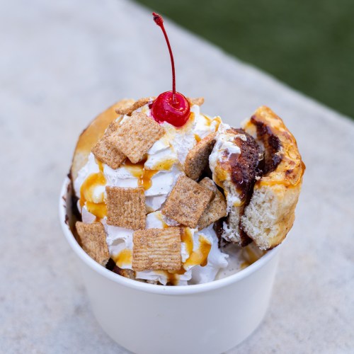 A dessert bowl with ice cream, whipped cream, a cherry on top, and cereal pieces, alongside a bite of cinnamon roll.