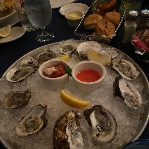 A platter of fresh oysters on ice, served with lemon wedges and dipping sauces, alongside rolls and a drink.