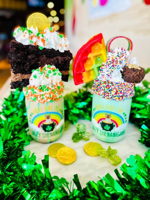 Two colorful milkshakes topped with whipped cream, sprinkles, and festive decorations, surrounded by green tinsel.