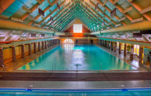 Indoor swimming pool with a high, vaulted ceiling and large windows, reflecting light on the water's surface.