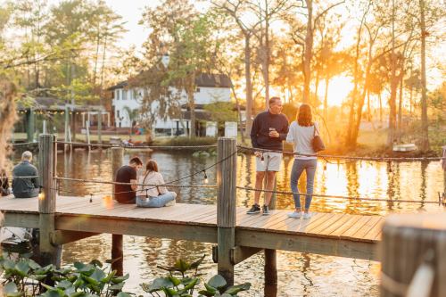 People enjoying a sunset on a wooden dock by a serene lake, with trees and a house in the background.