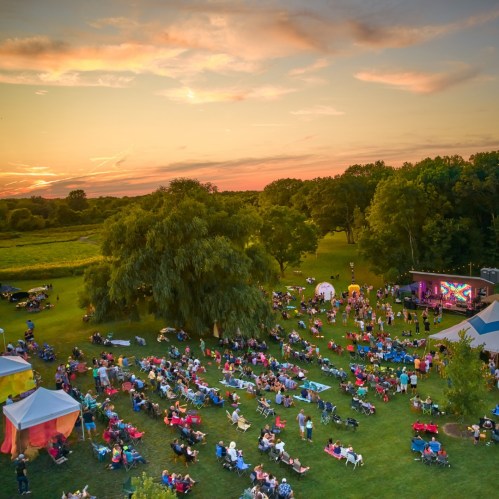 Aerial view of a lively outdoor concert at sunset, with a crowd gathered on the grass and colorful tents nearby.