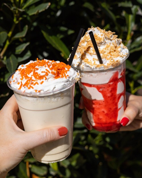 Two milkshakes in clear cups, one topped with orange sprinkles and whipped cream, the other with strawberry sauce and coconut.