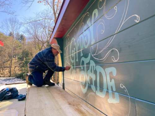 A person paints decorative lettering on a wooden wall outdoors, surrounded by trees and a clear sky.