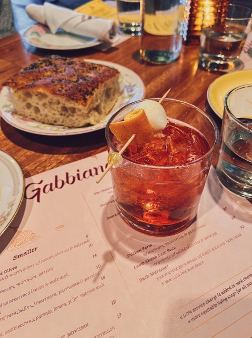 A cocktail with a garnish on a wooden table, alongside a menu and a piece of bread in the background.