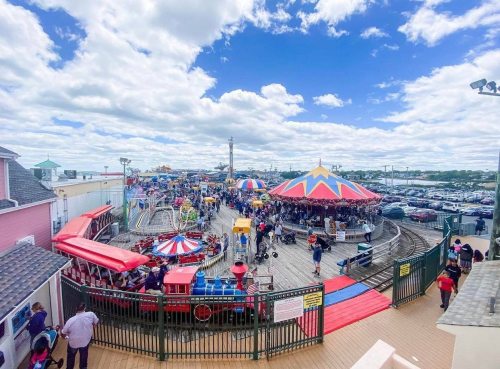 A vibrant amusement park scene with a carousel, rides, and a crowd under a blue sky with fluffy clouds.