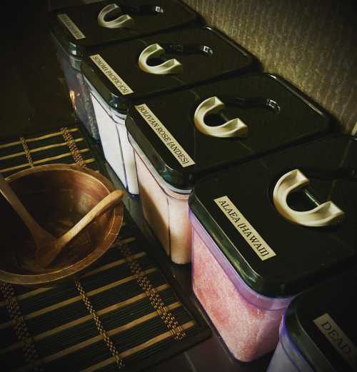 Containers of various salts labeled with names, alongside a wooden bowl and spoon on a woven mat.