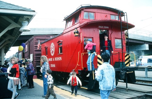 A red train caboose at a busy station with people boarding and disembarking, balloons in the background.