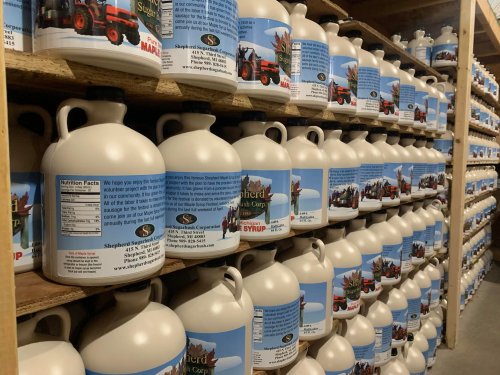 Rows of large, white syrup jugs with colorful labels stacked on wooden shelves in a storage area.