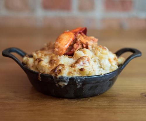 A cast-iron dish of creamy lobster mac and cheese, topped with a piece of lobster, on a wooden table.