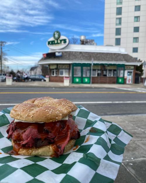 A close-up of a sesame seed bun sandwich filled with roast beef and sauce, with a restaurant in the background.