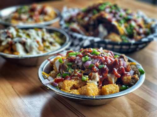 A close-up of a bowl of loaded tater tots topped with barbecue sauce, onions, and green peppers, with other dishes in the background.