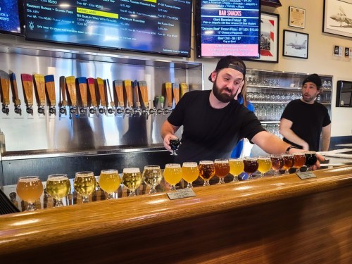A bartender serves a flight of colorful craft beers at a bar, with a menu displayed in the background.