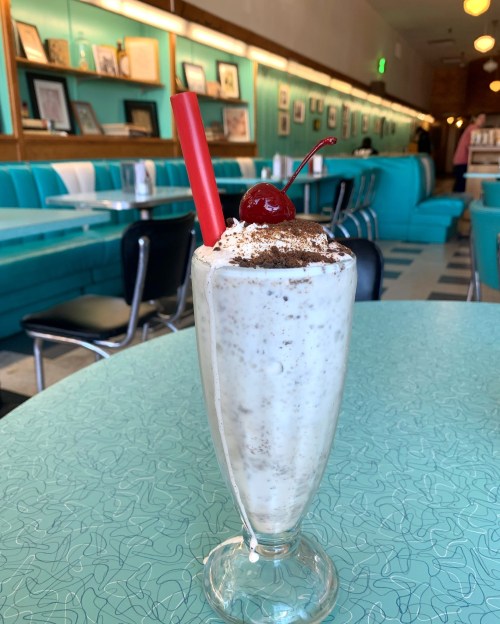 A tall glass of milkshake topped with whipped cream, chocolate sprinkles, a cherry, and a red straw on a retro diner table.