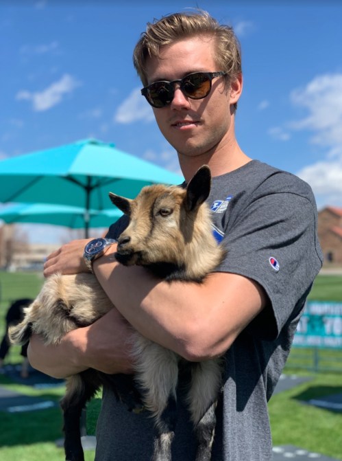 A young man in sunglasses holds a small goat, standing outdoors with blue skies and umbrellas in the background.