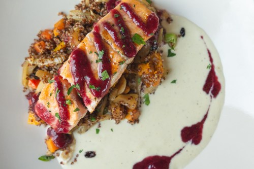 A plated dish featuring salmon with berry sauce, quinoa, roasted vegetables, and a creamy sauce on the side.