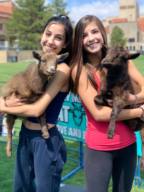 Two smiling young women hold baby goats in their arms, standing outdoors with a grassy background.