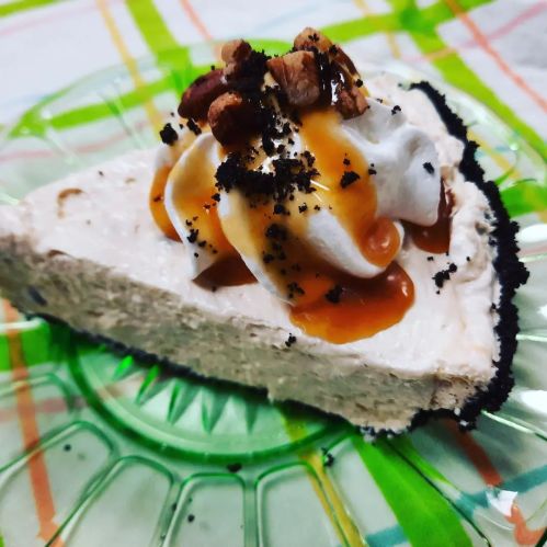 A slice of creamy dessert topped with whipped cream, caramel drizzle, and crushed nuts on a green glass plate.