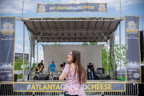 A woman in a floral top watches a band perform on stage at the Atlanta Grilled Cheese festival.