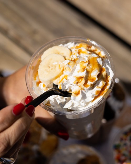 A hand holding a cup of dessert topped with whipped cream, banana slices, and caramel sauce, with a black straw.