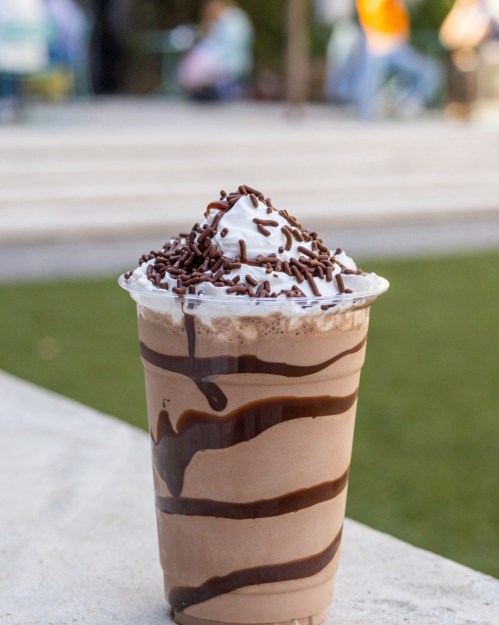 A chocolate milkshake topped with whipped cream and chocolate sprinkles, placed on a stone surface outdoors.