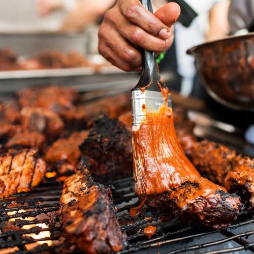 A hand brushes barbecue sauce onto grilled meat, with smoky, charred pieces visible on the grill.