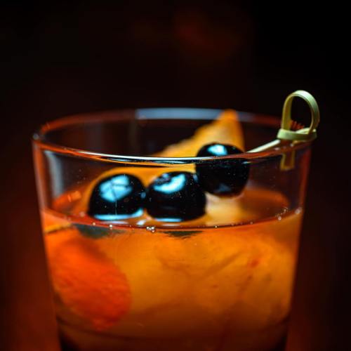 Close-up of a cocktail in a glass, featuring orange slices and three black olives on a skewer. Dark background.