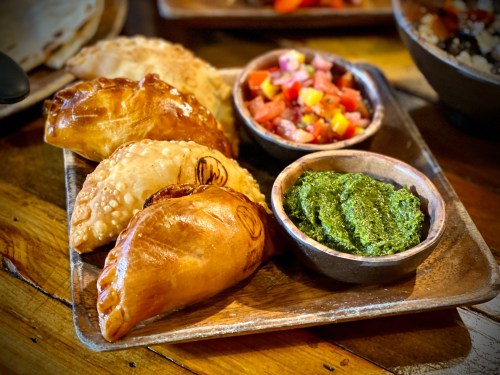A wooden platter with assorted empanadas, fresh salsa, and green sauce, set on a rustic table.