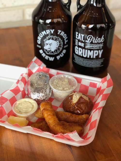 A takeout container with fried food, a baked potato, and sauces, alongside two growlers from Grumpy Troll Brew Pub.