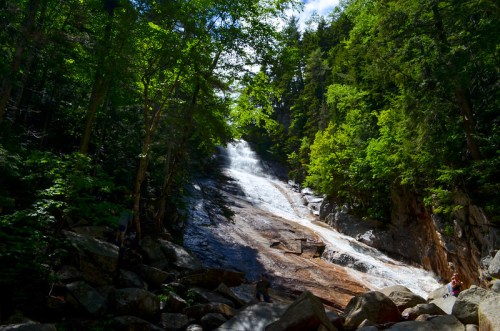 A serene waterfall cascades down rocky terrain, surrounded by lush green trees and a clear blue sky.