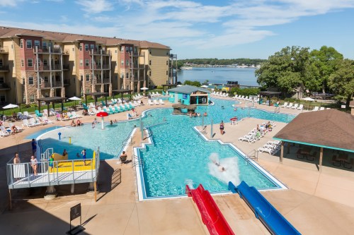 A vibrant pool area with water slides, lounge chairs, and people enjoying the sun by a lake and apartment buildings.