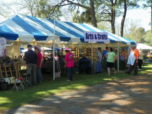 A colorful arts and crafts tent with people browsing and shopping in a park setting on a sunny day.