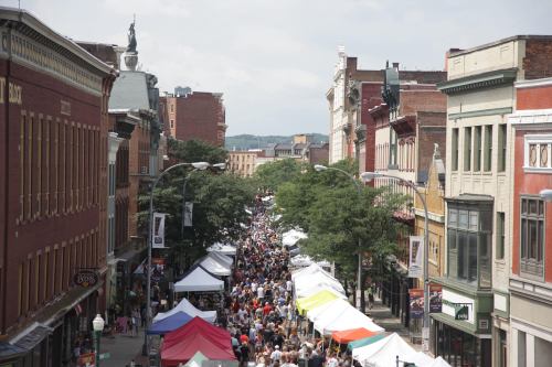 A bustling street fair with crowds and colorful tents lining the road, surrounded by historic buildings.