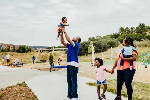 A father lifts his toddler in a park while a mother holds hands with another child, enjoying a sunny day outdoors.