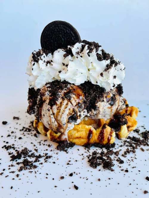 A delicious dessert featuring a waffle topped with ice cream, whipped cream, crushed cookies, and chocolate drizzle.