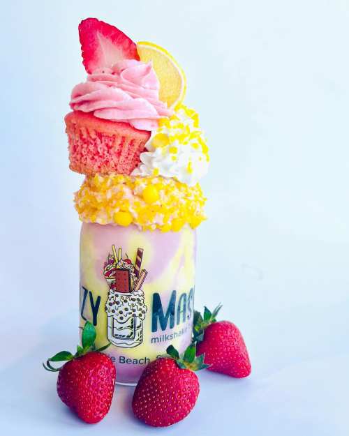 A colorful milkshake topped with whipped cream, a cupcake, strawberries, and lemon, served in a branded glass.