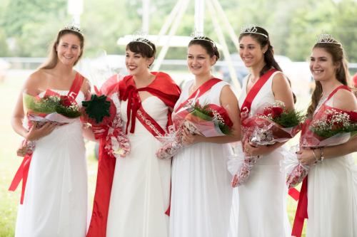 Five women in white dresses and tiaras, holding bouquets, smiling together at an outdoor event.