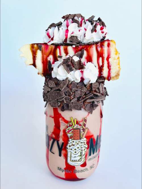 A decadent milkshake topped with whipped cream, chocolate shavings, and a slice of cake, drizzled with syrup.