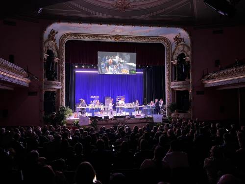 A large audience watches a performance on stage, with musicians and a screen displaying visuals in a grand theater.