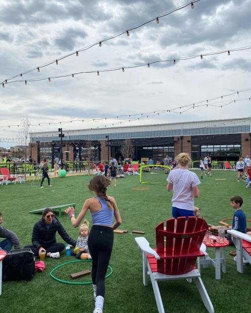 A lively outdoor space with people playing, children running, and adults relaxing on chairs under cloudy skies.
