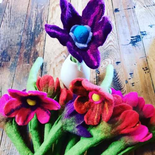Colorful felt flowers in a vase on a wooden surface, featuring vibrant petals and green stems.
