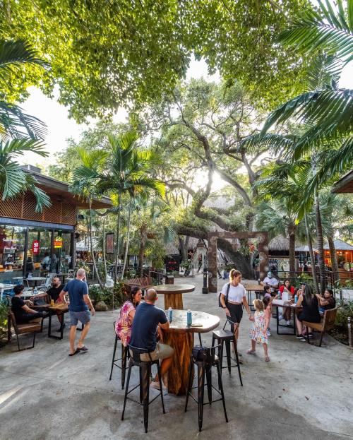 A lively outdoor space with people socializing around tables, surrounded by lush greenery and tropical trees.