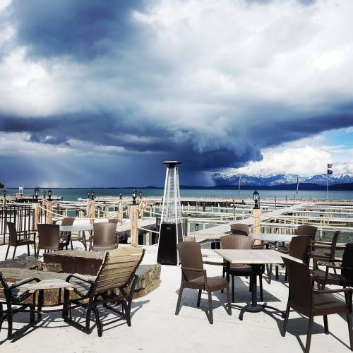 A lakeside patio with empty tables and chairs, under a dramatic sky with dark clouds and distant snow-capped mountains.