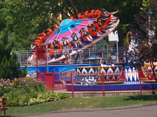 A colorful amusement park ride spins with excited riders, surrounded by greenery and vibrant flowers.
