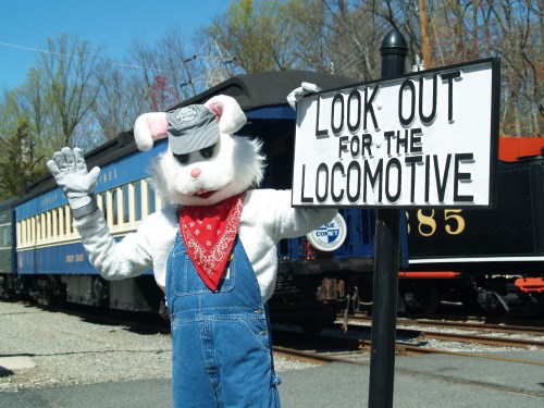 A person in a rabbit costume waves beside a sign that reads "LOOK OUT FOR THE LOCOMOTIVE" near a train.